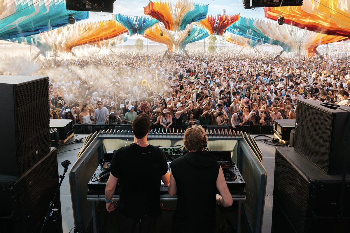 DJ duo at the decks facing a festival crowd beneath a canopy of colored fabric